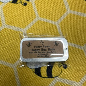 May include: A small, rectangular tin of Happy Bee Balm, made by Honey Farms, is displayed on a yellow background with a honeycomb pattern and bee illustrations. The balm is made with beeswax, shea butter, and jojoba oil, with a net weight of 0.20 ounces.