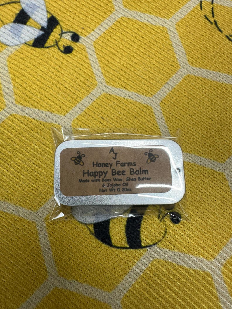 May include: A small, rectangular tin of Happy Bee Balm, made by Honey Farms, is displayed on a yellow background with a honeycomb pattern and bee illustrations. The balm is made with beeswax, shea butter, and jojoba oil, with a net weight of 0.20 ounces.