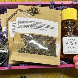 May include: A pink gift box filled with purple tissue paper contains a small glass jar of honey with a bear-shaped label that reads "Honey A.J. Farms Raw Honey".  The box also contains a small brown paper bag with a label that reads "Lights Out - Sleepy Blend WI 2.5g" and a silver heart-shaped tea infuser.
