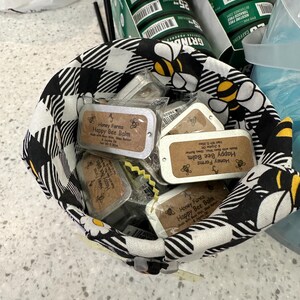 May include: A black and white checkered fabric basket filled with small rectangular tins of "Honey Farms Happy Bee Balm." The basket has a bee and daisy pattern. Behind the basket are green containers with the word "GRIND" and the text "CAFFEINE".