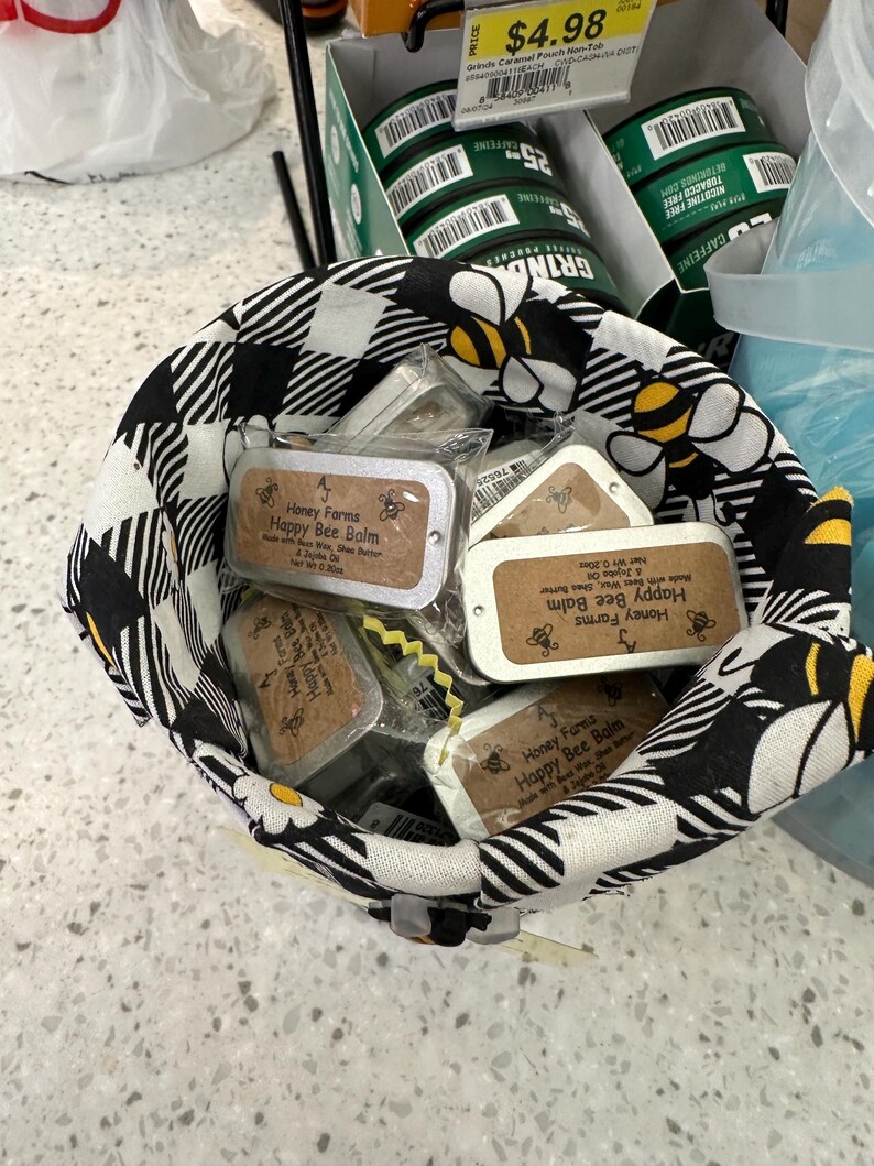 May include: A black and white checkered fabric basket filled with small rectangular tins of "Honey Farms Happy Bee Balm." The basket has a bee and daisy pattern. Behind the basket are green containers with the word "GRIND" and the text "CAFFEINE".
