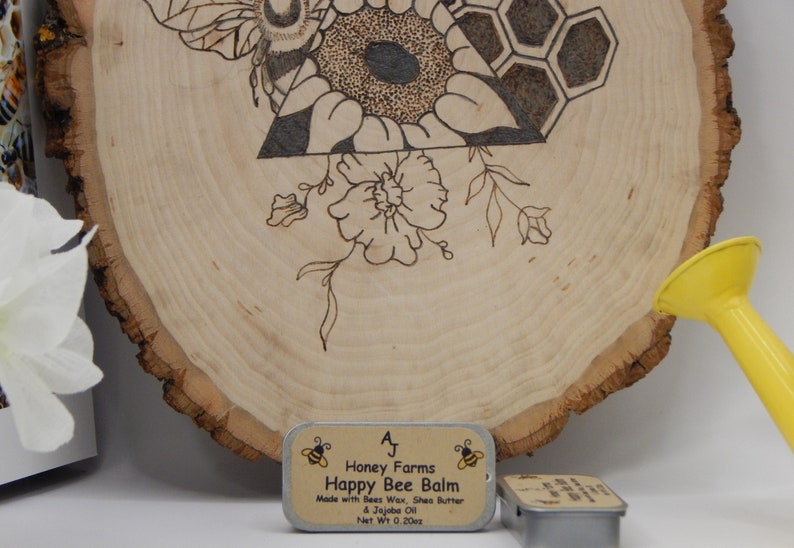 May include: A wooden slice with a burned bee and floral design, with a small tin of Happy Bee Balm. The balm is made with beeswax, shea butter, and jojoba oil, net weight 0.20oz.