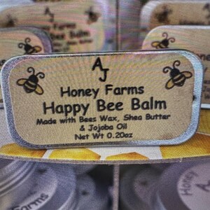 May include: A close-up of a rectangular label for "Happy Bee Balm" from "Honey Farms." The label features bee illustrations and text indicating the balm is made with beeswax, shea butter, and jojoba oil. Net weight is 0.20oz.