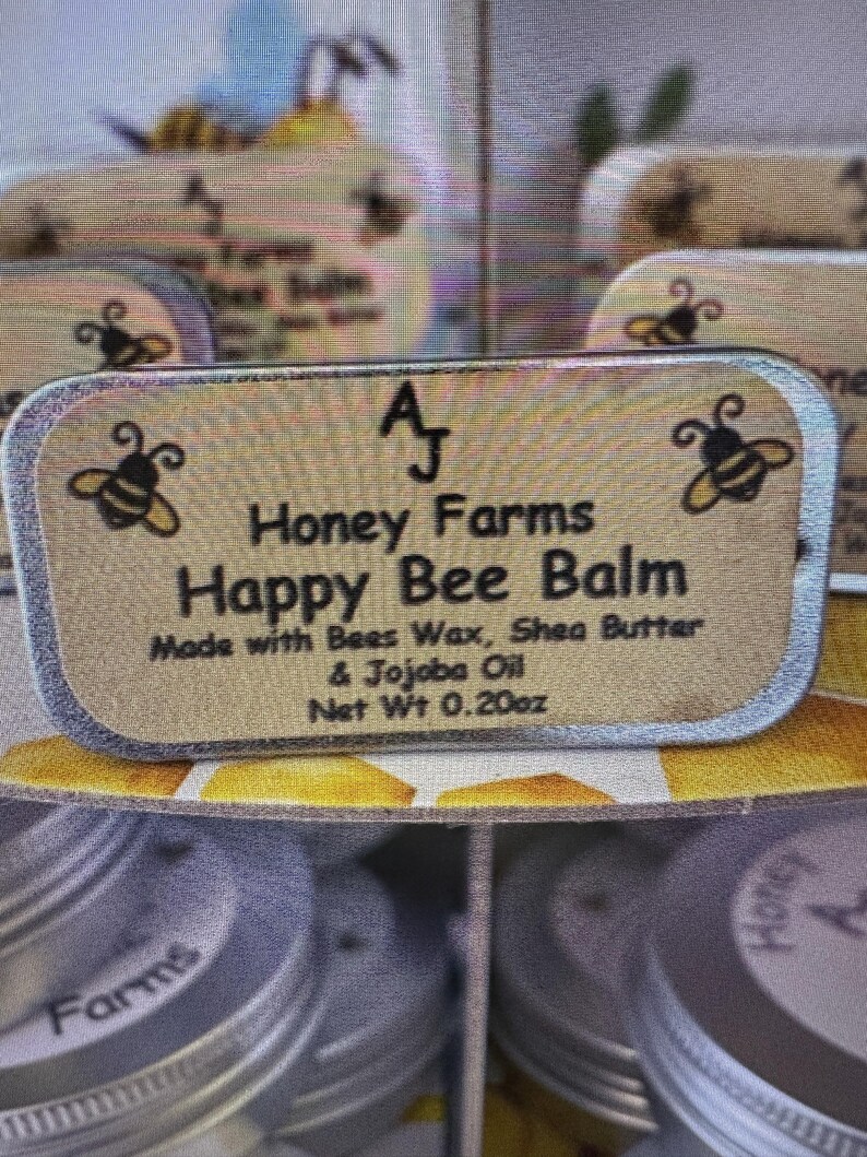May include: A close-up of a rectangular label for "Happy Bee Balm" from "Honey Farms." The label features bee illustrations and text indicating the balm is made with beeswax, shea butter, and jojoba oil. Net weight is 0.20oz.