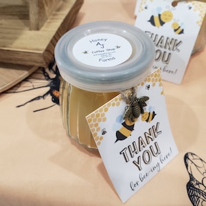 May include: A small glass jar filled with honey, topped with a clear lid. The jar has a label that reads "Honey AJ Coffee Shop Farms." A "Thank You" tag with bee graphics is attached. The tag reads "Thank You for bee-ing here!"