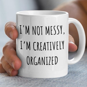 May include: White ceramic mug with the text "I'M NOT MESSY, I'M CREATIVELY ORGANIZED" in black. The mug has a curved handle and is held by a person. The mug is a standard size for coffee or tea.