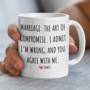 May include: White ceramic coffee mug with black text that reads "Marriage: The art of compromise. I admit I'm wrong, and you agree with me." A small red heart with the name "James" is below the text.