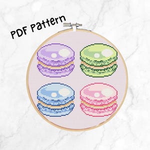 Op de afbeelding: Kruissteekpatroon voor vier kleurrijke macarons. Het patroon wordt weergegeven in een houten borduurring. De macarons zijn paars, groen, blauw en roze.