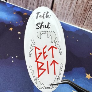 May include: White oval sticker with the text "Talk Shit" above "Get Bit" in red, with a stylized mouth graphic. The sticker is held by tweezers against a blue background with a starry design.