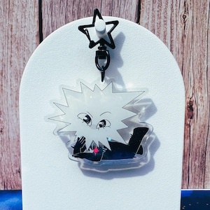 May include: A clear acrylic keychain featuring a cartoon character with spiky white hair and a black and blue outfit. The keychain has a black star-shaped clasp. The character is lying down with a red star on its chest.