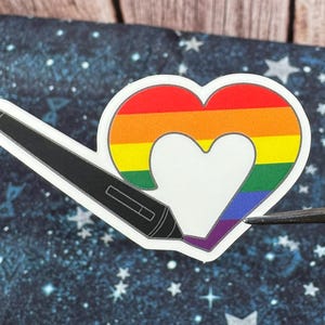 Puede incluir: Una pegatina troquelada que muestra un lápiz óptico digital negro dibujando una forma de corazón. El corazón está relleno con rayas horizontales de arcoíris y tiene un pequeño corazón blanco recortado en el centro. La pegatina tiene un borde blanco.