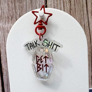 May include: A clear acrylic keychain with the text "TALK SHIT GET BIT" in black and red. The keychain has a red star-shaped clip and a design of a skull with teeth. The keychain is hanging on a white surface.