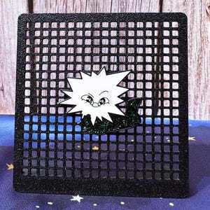 May include: A white cartoon sunburst pin with a smiling face, set against a black grid. The pin is attached to a dark green, glittery base. The background features a blue and black gradient.