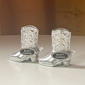 May include: A pair of silver cowboy boot salt and pepper shakers with black labels that read "Banff Canada".