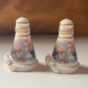 Può includere: Un paio di saliere e pepiere in ceramica. Ogni shaker presenta una scena di faro con un edificio dal tetto rosso, sullo sfondo di un cielo e di un oceano colorati. Gli shaker hanno finiture dorate e poggiano su una base a forma di onda.
