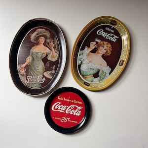 Puede incluir: Tres bandejas vintage de Coca-Cola. Dos bandejas ovaladas muestran a una mujer sosteniendo un vaso de Coca-Cola. Una bandeja es negra con un borde blanco y la otra es amarilla con un borde verde. La tercera bandeja es redonda y roja con un borde negro. Dice "Take home a carton Coca-Cola 6 bottles 25¢ plus deposit".