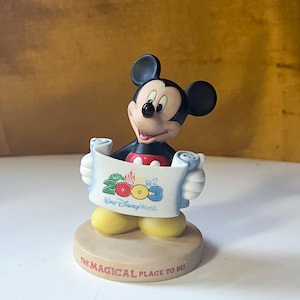 May include: A Mickey Mouse figurine holding a scroll that reads "2003 Walt Disney World." The figurine has black ears, a red shirt, and yellow shoes. The base reads "THE MAGICAL PLACE TO BE!"