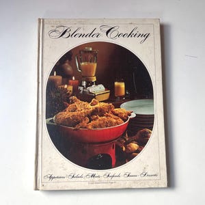 以下が含まれることがあります： クリーム色の表紙の「Blender Cooking」というタイトルのヴィンテージ料理本。表紙には、ミキサー、食べ物、ろうそくの円形画像が掲載されています。本の表紙には、前菜、サラダ、肉、シーフード、ソース、デザートなどの食品カテゴリーも記載されています。
