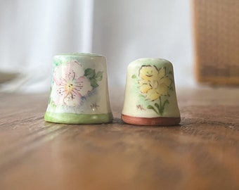 May include: Two ceramic thimbles with floral designs. The thimble on the left has a pink flower with green leaves. The thimble on the right has a yellow flower with green leaves.