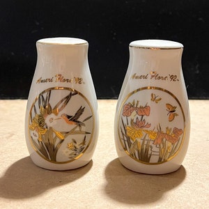 May include: A pair of white ceramic salt and pepper shakers with gold trim. The shakers feature a floral design with a hummingbird on one and butterflies on the other. The text "Ameri Flora '92" is printed on both shakers.