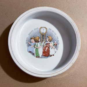May include: White ceramic dish with a gold rim featuring a nursery rhyme illustration. The image depicts children gathered around a grandfather clock, with the text "Hickory, Dickory, Dock" and "The mouse ran up the clock."