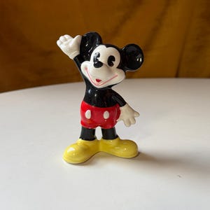 Puede incluir: Una figura de cer&aacute;mica de Mickey Mouse, un personaje de dibujos animados cl&aacute;sico. Mickey est&aacute; representado con orejas negras, cuerpo negro, pantalones cortos rojos, zapatos amarillos y guantes blancos. Est&aacute; posando con un brazo levantado.
