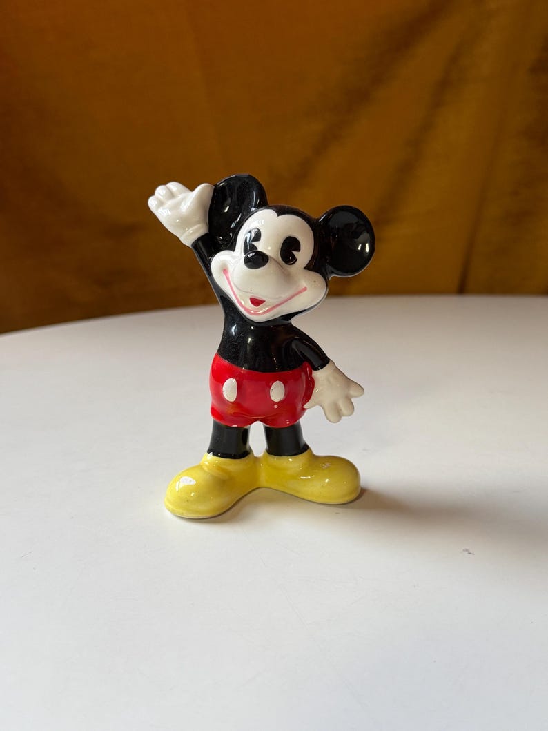Puede incluir: Una figura de cer&aacute;mica de Mickey Mouse, un personaje de dibujos animados cl&aacute;sico. Mickey est&aacute; representado con orejas negras, cuerpo negro, pantalones cortos rojos, zapatos amarillos y guantes blancos. Est&aacute; posando con un brazo levantado.