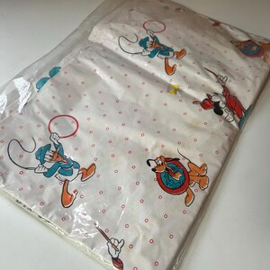Puede incluir: Un juego de s&aacute;banas blancas empaquetado con personajes de dibujos animados como el Pato Donald, Mickey Mouse y Pluto. Las s&aacute;banas tienen un patr&oacute;n repetitivo de puntos rojos. Los personajes est&aacute;n en varias poses, algunos sosteniendo aros.