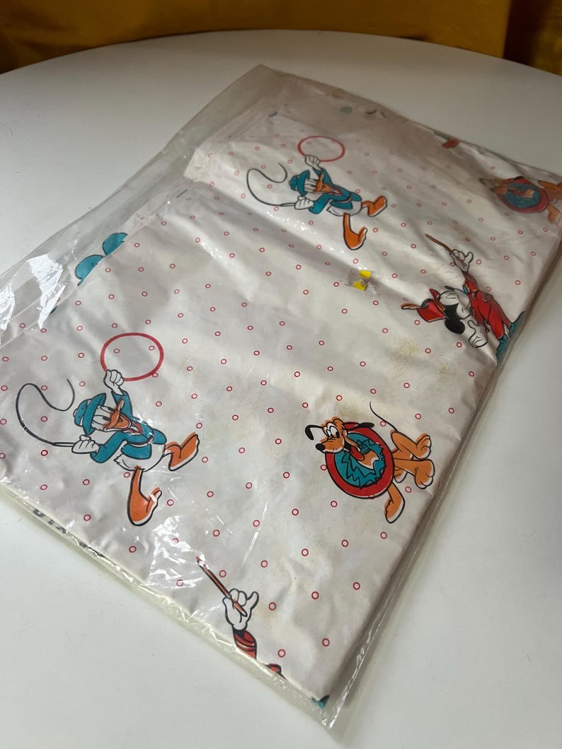 Puede incluir: Un juego de s&aacute;banas blancas empaquetado con personajes de dibujos animados como el Pato Donald, Mickey Mouse y Pluto. Las s&aacute;banas tienen un patr&oacute;n repetitivo de puntos rojos. Los personajes est&aacute;n en varias poses, algunos sosteniendo aros.