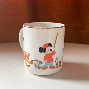 May include: White ceramic mug with a gold rim featuring cartoon characters. Mickey Mouse is in the center, wearing a red jacket and hat, holding a wooden stick. Other characters include Pluto and a rabbit. The mug has a curved handle.
