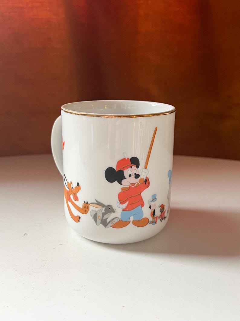 May include: White ceramic mug with a gold rim featuring cartoon characters. Mickey Mouse is in the center, wearing a red jacket and hat, holding a wooden stick. Other characters include Pluto and a rabbit. The mug has a curved handle.