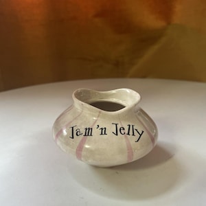 Op de afbeelding: Een kleine, witte keramische pot met roze strepen en de tekst "Jam 'n Jelly" erop.