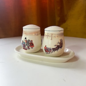 Könnte beinhalten: Ein Set aus zwei Salz- und Pfefferstreuern aus Keramik mit einem Wintermotiv. Die Streuer sind weiß mit grünem Rand und zeigen eine Winterszene mit Kindern, die im Schnee spielen, und einem Schlitten, der von Rentieren gezogen wird.
