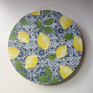 May include: Round decorative serving tray with a lemon and leaf design. The tray features a blue and white tile pattern background with yellow lemons and green leaves. The tray is circular.