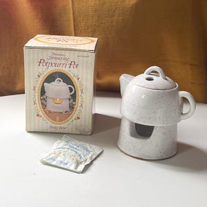 Puede incluir: Una olla de popurrí de cerámica blanca con motas rosas, una caja a juego y una bolsita. La caja dice "Traditional Simmering Potpourri Pot" con una imagen de la olla. La olla tiene forma de tetera.