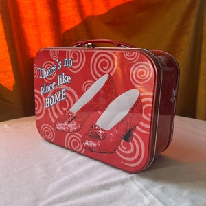 Puede incluir: Fiambrera metálica roja con asa, con un diseño de zapatos rojos con lentejuelas y el texto "There's no place like HOME". La fiambrera tiene un interior blanco y un exterior rojo con un patrón de remolino.