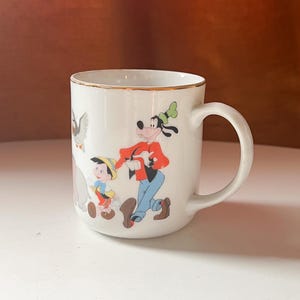 May include: White ceramic mug with a gold rim, decorated with Disney characters such as Goofy and Pinocchio. The mug has a curved handle and is set against a neutral background. The design is colourful and whimsical, ideal for a Disney fan.