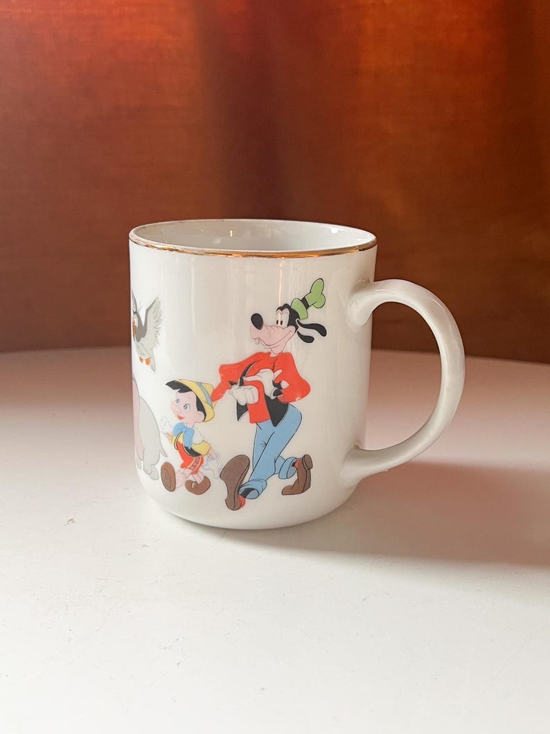 May include: White ceramic mug with a gold rim, decorated with Disney characters such as Goofy and Pinocchio. The mug has a curved handle and is set against a neutral background. The design is colourful and whimsical, ideal for a Disney fan.