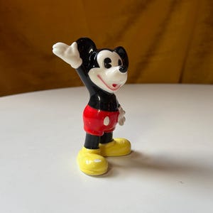 Puede incluir: Figura de cer&aacute;mica de Mickey Mouse, un personaje de dibujos animados cl&aacute;sico. Mickey tiene orejas negras, cuerpo negro, pantalones cortos rojos, zapatos amarillos y guantes blancos. Est&aacute; saludando con la mano derecha. La figura est&aacute; sobre una superficie blanca.