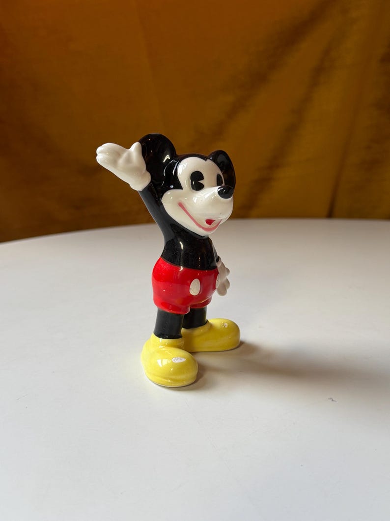 Puede incluir: Figura de cer&aacute;mica de Mickey Mouse, un personaje de dibujos animados cl&aacute;sico. Mickey tiene orejas negras, cuerpo negro, pantalones cortos rojos, zapatos amarillos y guantes blancos. Est&aacute; saludando con la mano derecha. La figura est&aacute; sobre una superficie blanca.