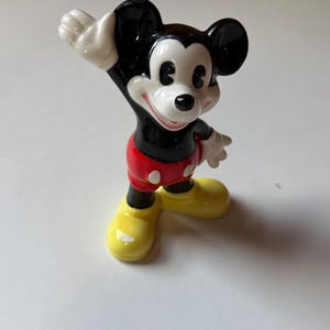 Puede incluir: Una figura de cer&aacute;mica de Mickey Mouse, un personaje cl&aacute;sico de Disney. Mickey tiene orejas negras, cuerpo negro, pantalones cortos rojos con botones blancos, zapatos amarillos y guantes blancos. Est&aacute; saludando con una sonrisa.