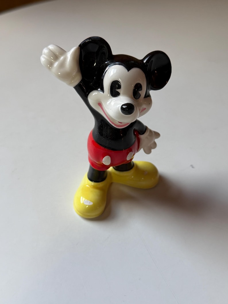 Puede incluir: Una figura de cer&aacute;mica de Mickey Mouse, un personaje cl&aacute;sico de Disney. Mickey tiene orejas negras, cuerpo negro, pantalones cortos rojos con botones blancos, zapatos amarillos y guantes blancos. Est&aacute; saludando con una sonrisa.