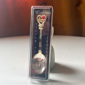 Może przedstawiać: Srebrna łyżeczka z pozłacanym serduszkiem z napisem "I Love New York" na miseczce. Łyżeczka jest zapakowana w przezroczyste plastikowe pudełko z czarnym tłem i nadrukiem "Watsons Silver Plated Spoon".