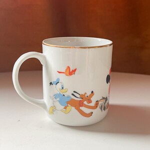 May include: White ceramic mug with a gold rim, depicting classic cartoon characters such as Donald Duck and Pluto. The mug has a curved handle and is decorated with colourful illustrations of the characters in action.