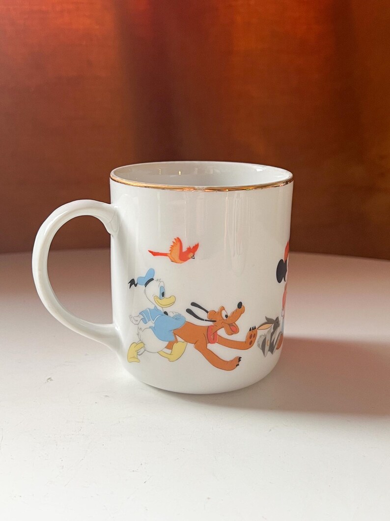 May include: White ceramic mug with a gold rim, depicting classic cartoon characters such as Donald Duck and Pluto. The mug has a curved handle and is decorated with colourful illustrations of the characters in action.