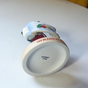 May include: A ceramic Mickey Mouse figurine, dressed in a red jacket and black shorts, holding a banner with "The Magical" written on it. The base is off-white with the Disney logo and "China" printed.