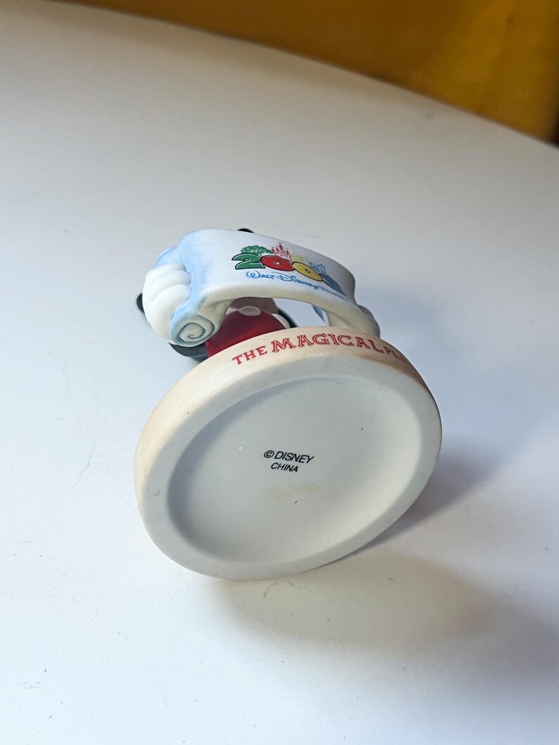 May include: A ceramic Mickey Mouse figurine, dressed in a red jacket and black shorts, holding a banner with "The Magical" written on it. The base is off-white with the Disney logo and "China" printed.