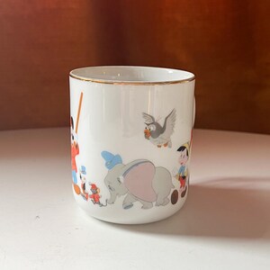 May include: White ceramic mug with a gold rim, featuring colourful illustrations of Disney characters. The design includes Dumbo the elephant, Pinocchio, and other classic characters. The mug is set against a white surface and a brown background.