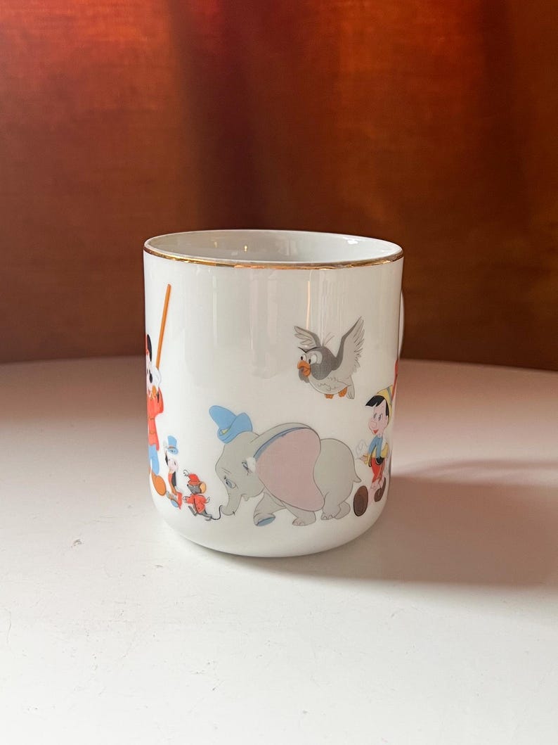 May include: White ceramic mug with a gold rim, featuring colourful illustrations of Disney characters. The design includes Dumbo the elephant, Pinocchio, and other classic characters. The mug is set against a white surface and a brown background.