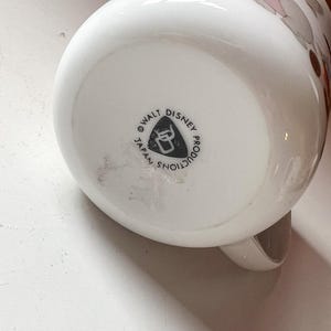 May include: White ceramic mug with a cartoon illustration. The base of the mug features the text "WALT DISNEY PRODUCTIONS JAPAN SHOW". The mug has a handle and a rounded shape. The mug is likely vintage.